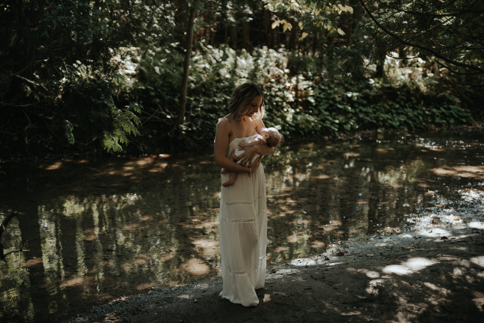 New mother carrying newborn daughter by a river in a forest for newborn portraits