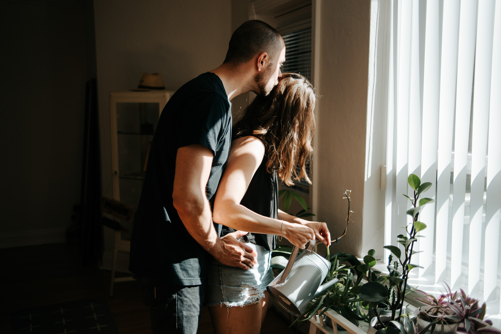 Guy hugging girl from behind as she waters plants in apartment