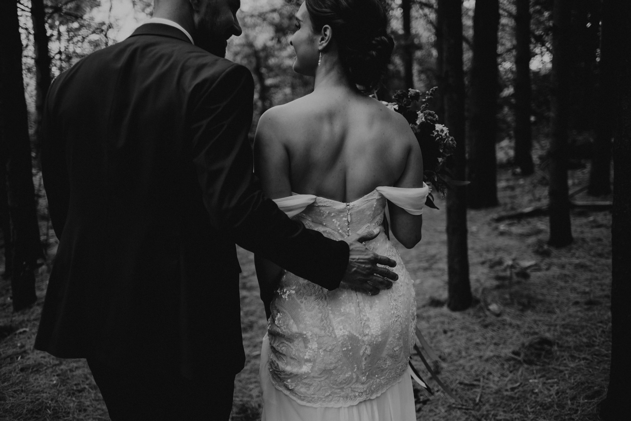 Groom's hand on back of bride's dress as they walk in forest