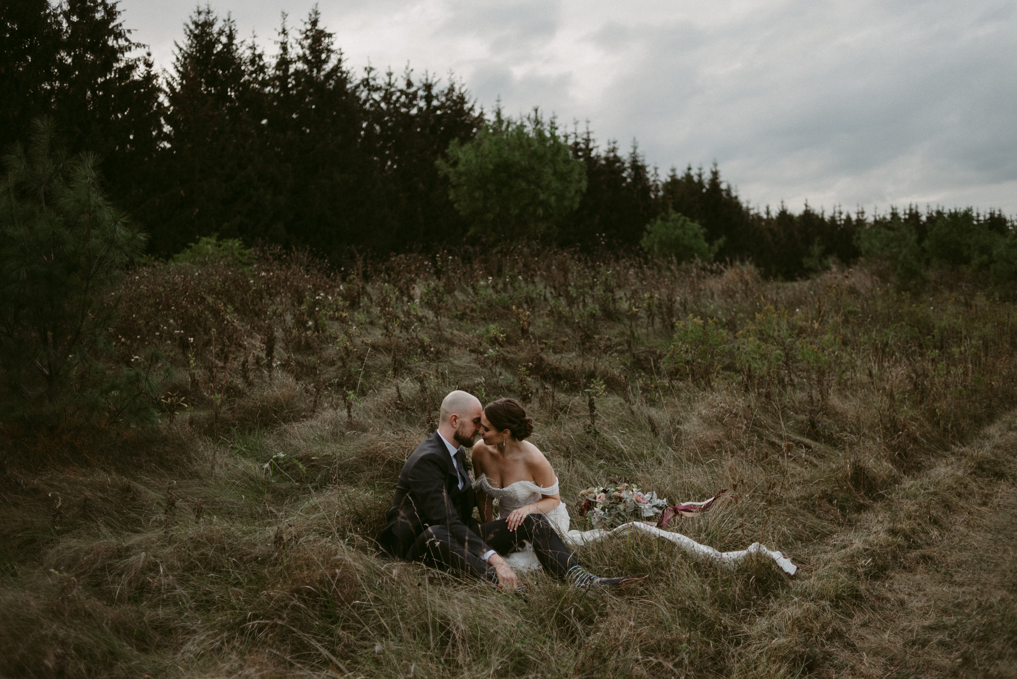 Bride and groom sitting in tall grass beside forest