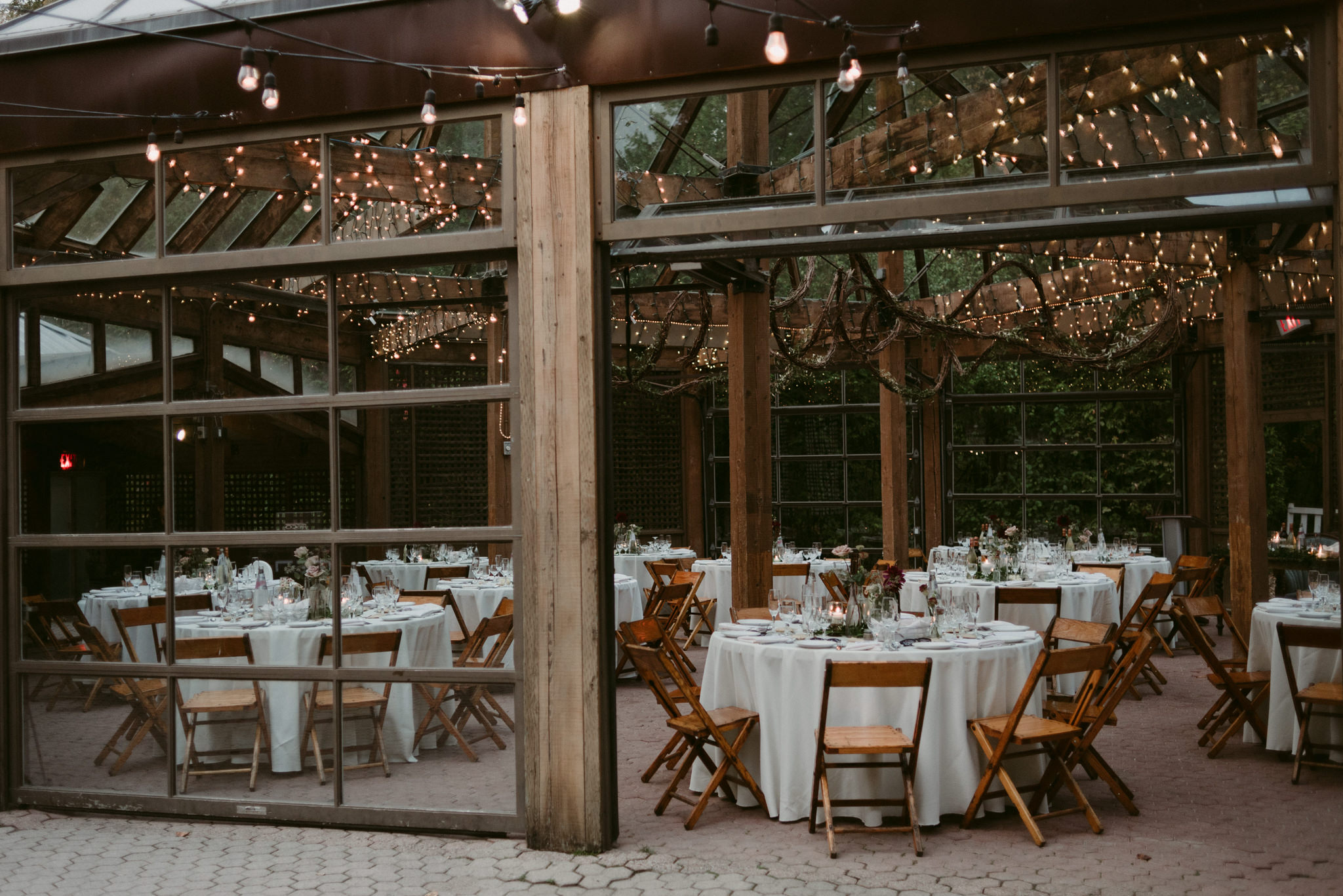 Glass room wedding reception set up with simple white linen round tables and twinkling lights