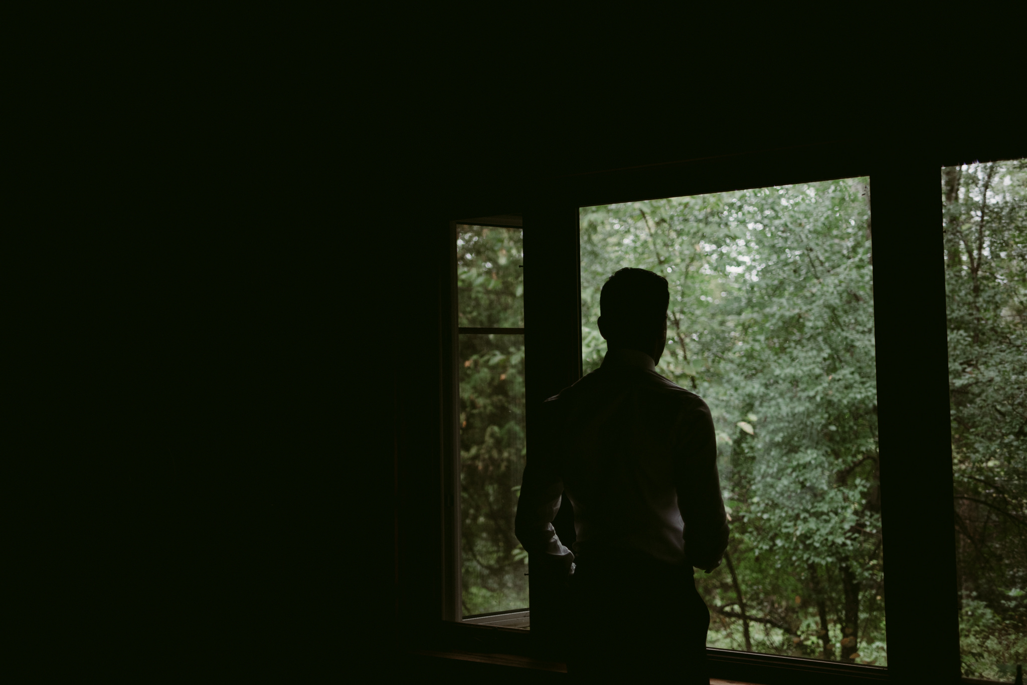 Groom standing and looking out window at forest in cottage