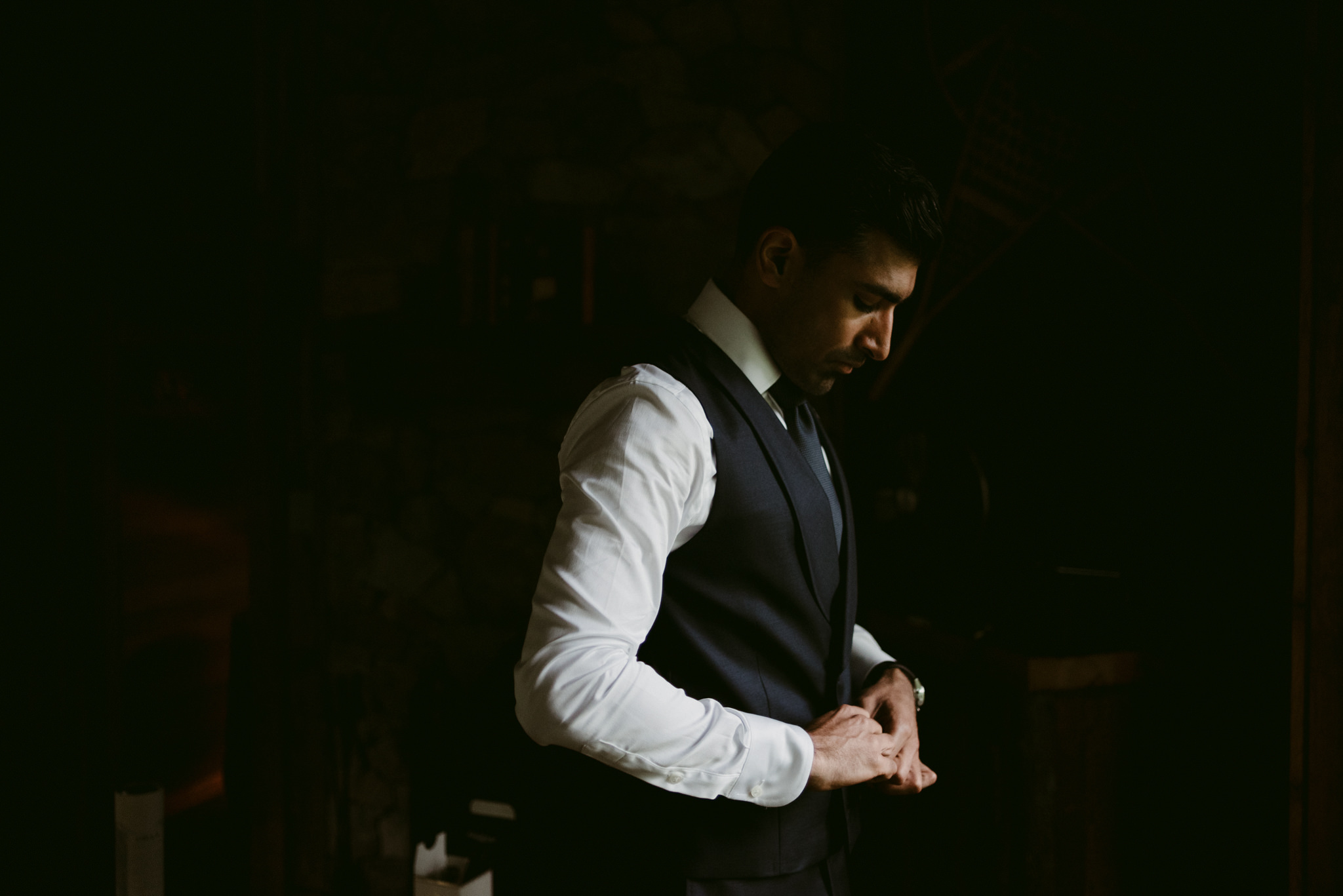 groom getting dressed and buttoning up vest in dark cottage