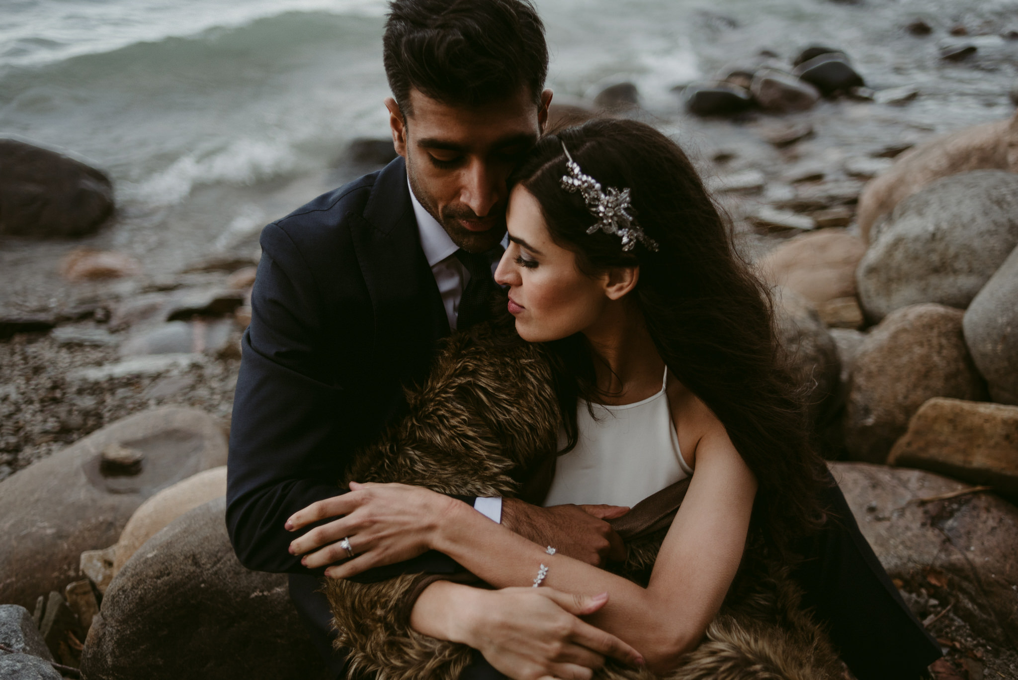 intimate portrait of bride and groom sitting on rocks by water