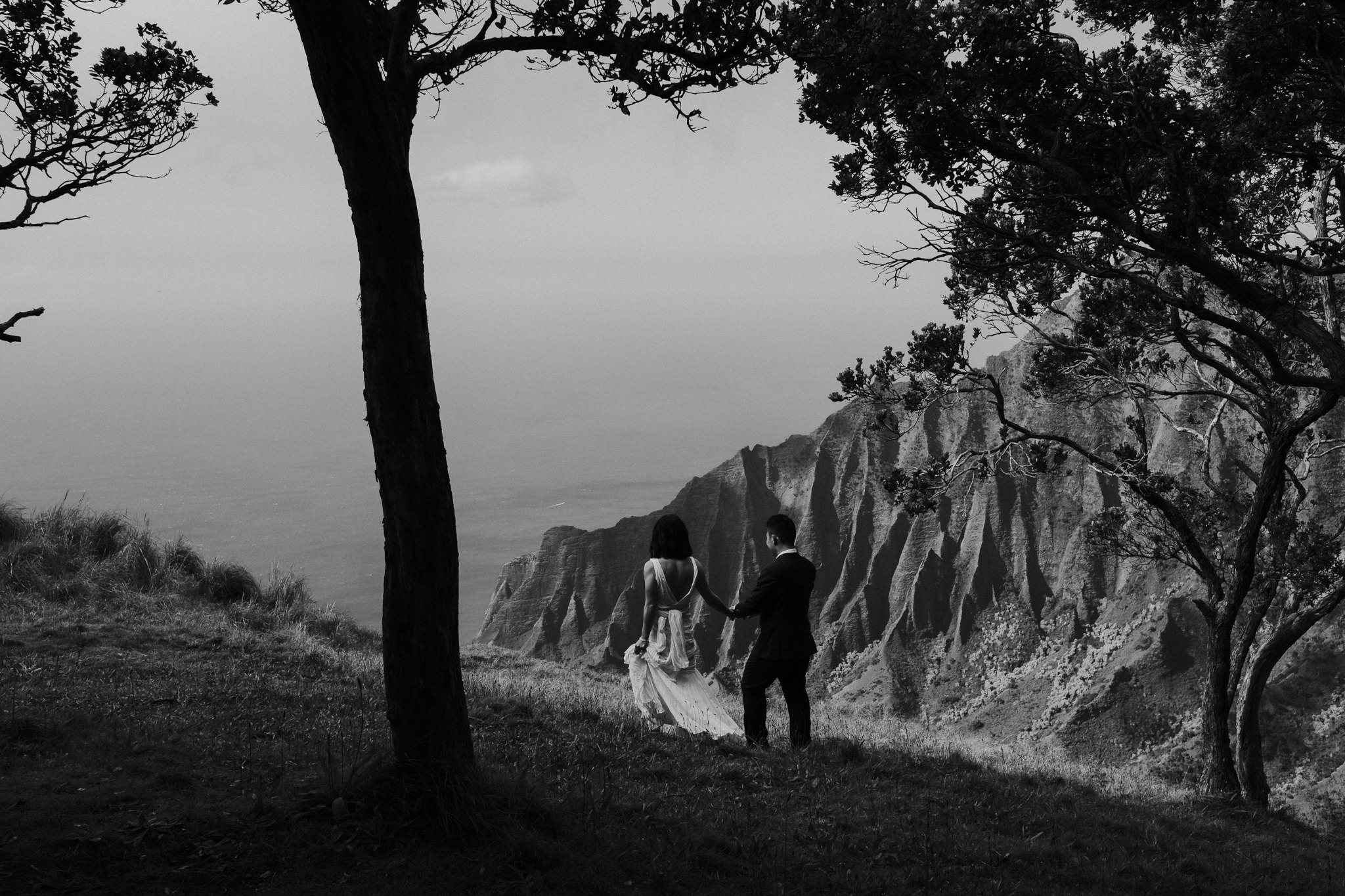 bride and groom walking on cliff edge with view of na pali coast in kauai