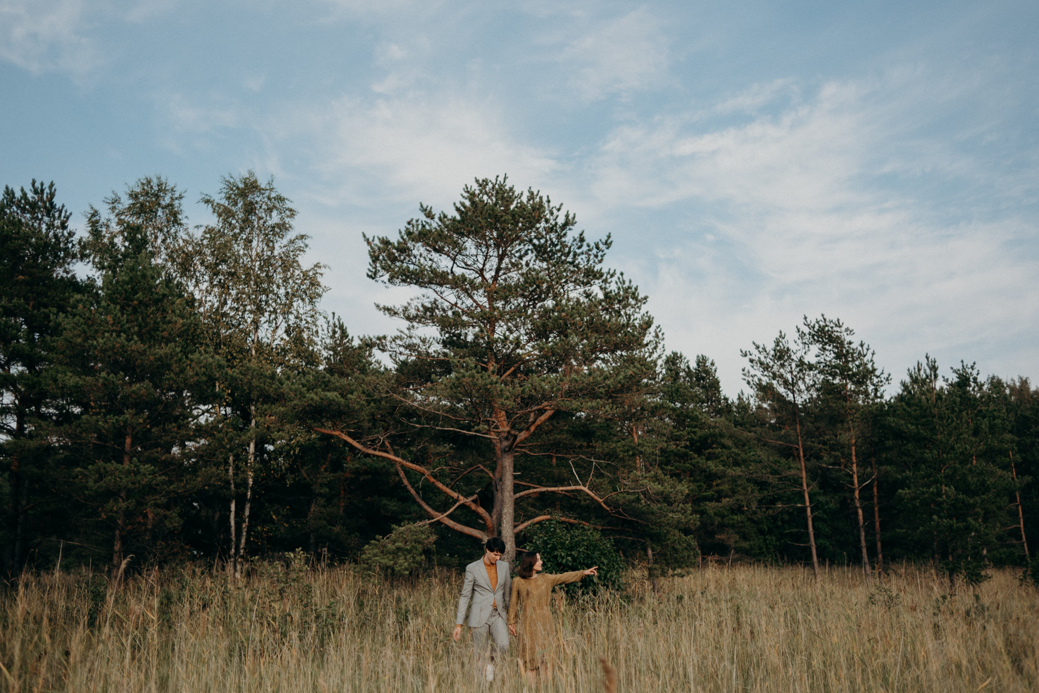 man in suit and woman in dress walking in tall grass near forest in Helsinki