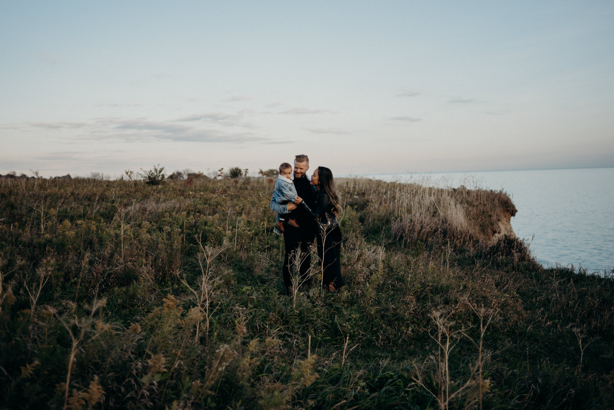 young family with baby boy standing by Lake Ontario at sunset