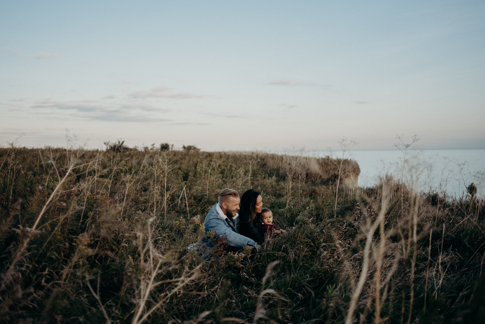 young couple cuddling with baby boy in the grass at sunset