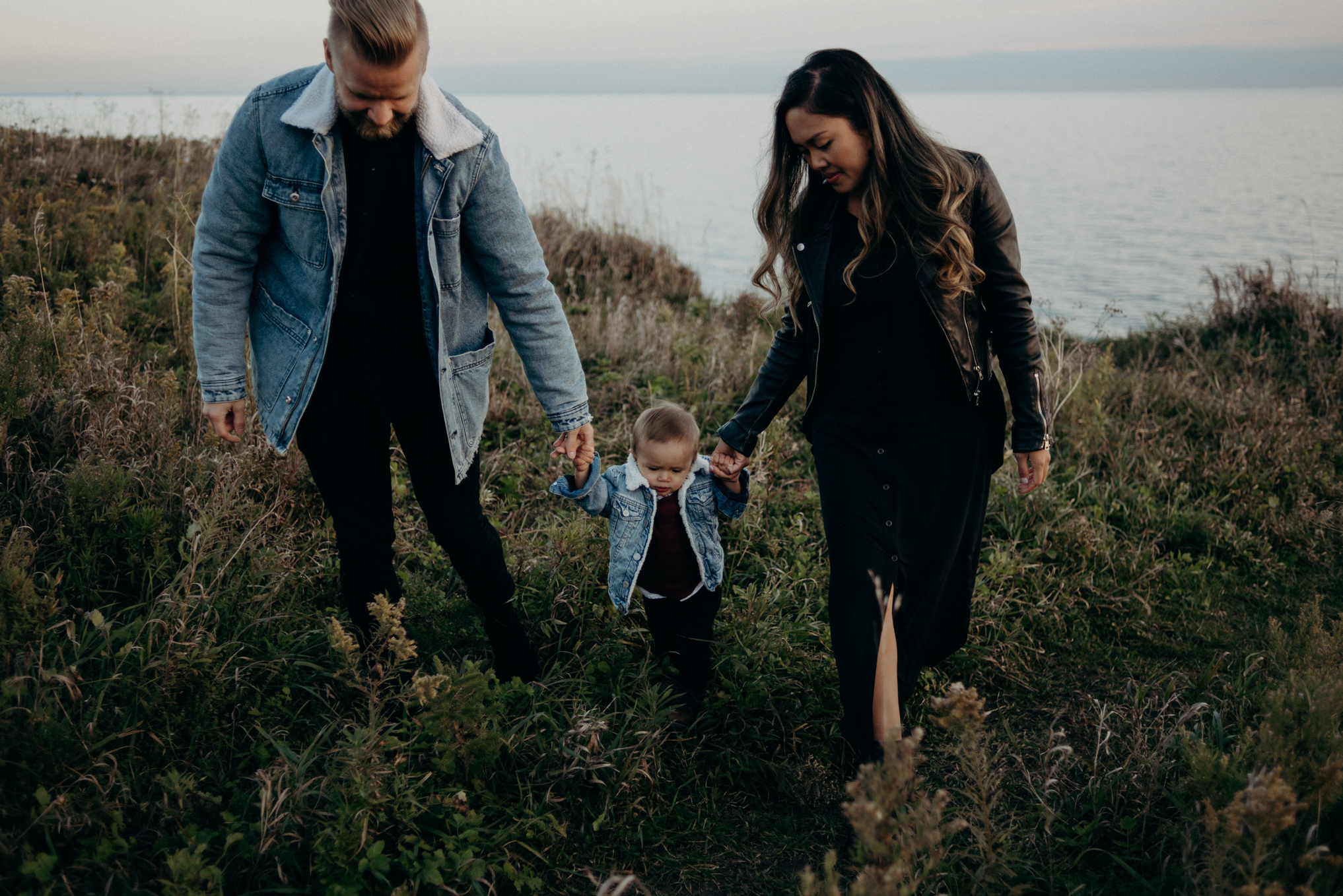 mixed baby boy trying to walk outside in grass while holding mom and dads hands