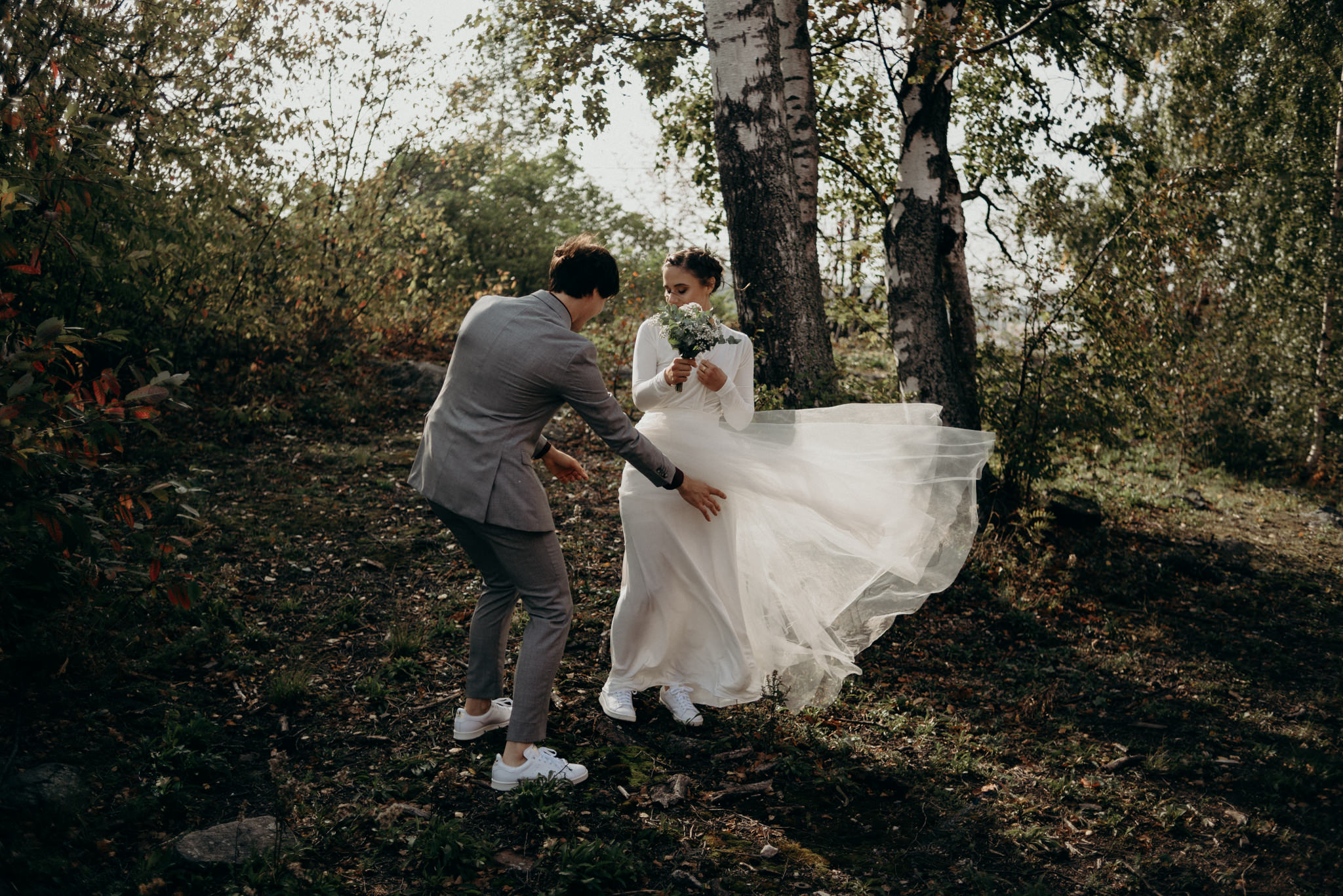 groom adjusting brides dress that is blowing in wind as they stand among trees in the sunlight