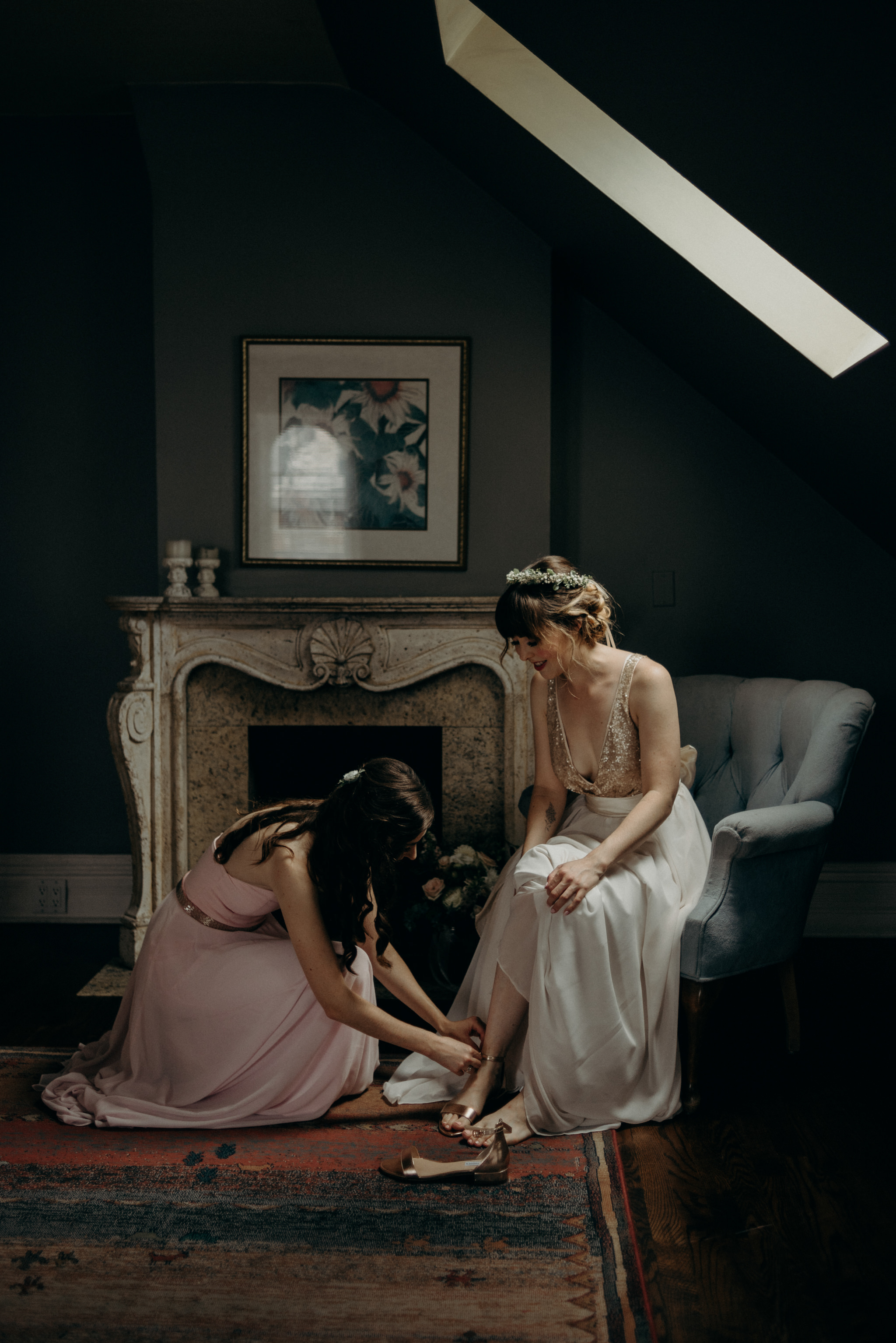 bridesmaid helping bride into shoes as she sits on chair
