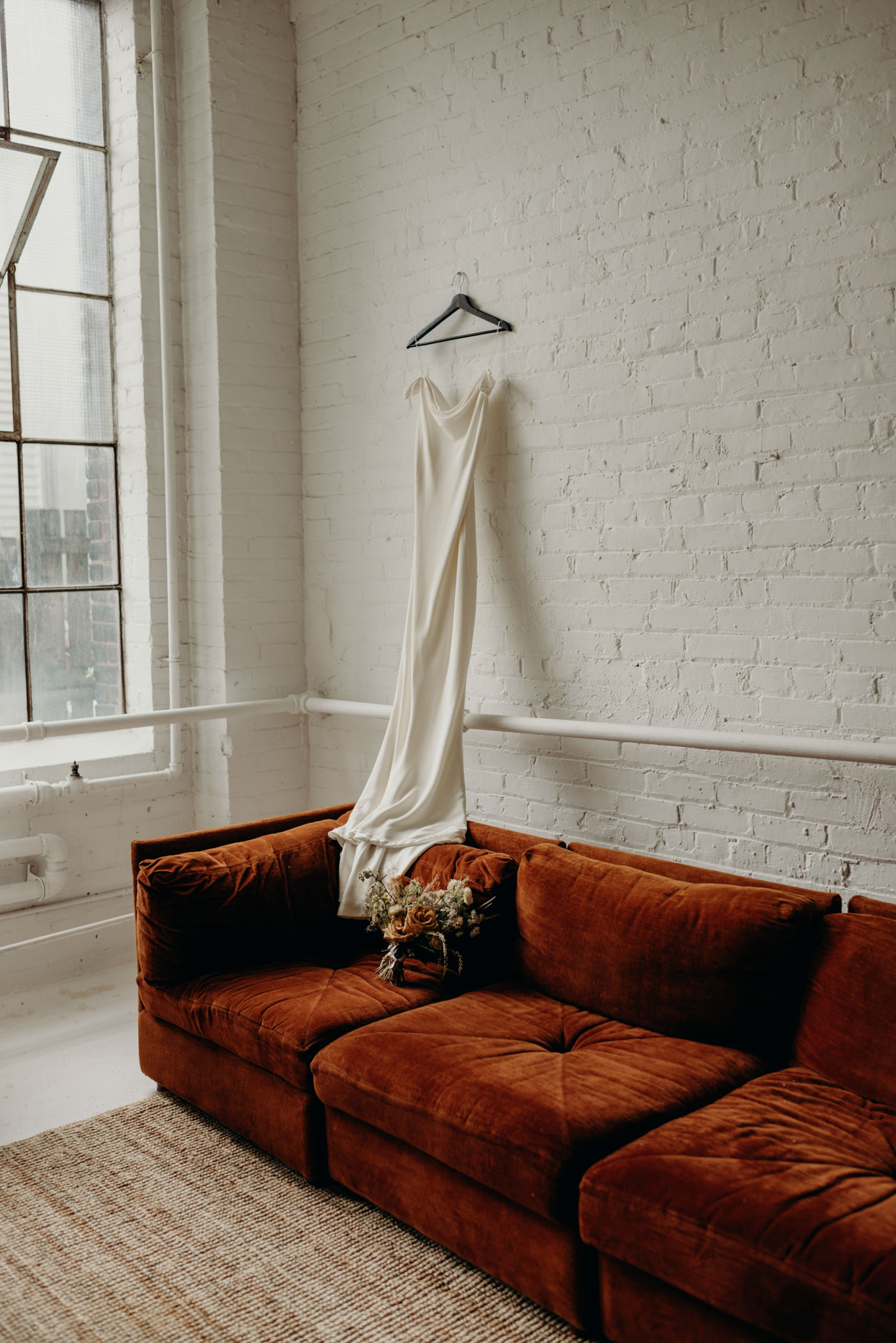 wedding dress hanging on white brick wall with flowers on velvet couch