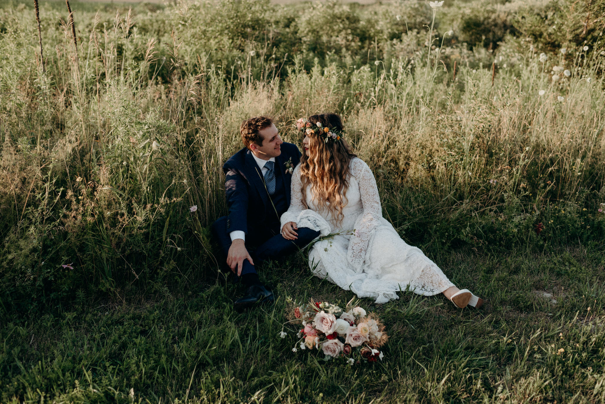 couple sitting in tall grass surrounded by flowers at sunset